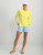 HUK Huk Pursuit Graphic Huk Box, woman wearing yellow long-sleeve athletic shirt, light blue shorts, and white cap, smiling and standing against a white background.