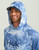 HUK Pursuit Mossy Oak Printed Hoodie, a man wearing a blue camouflage hoodie with a hood and a cap underneath, featuring performance fishing branding and Mossy Oak logo.