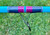 Nick's Custom Rods 2025 Fall Nick's Custom Rods - CTS 1205 Spinning, close-up of a colorful spinning rod with pink, blue, and purple striped design on the handle and a metallic guide against a grassy background.