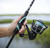 Toadfish Carbon II Spinning Combos, close-up view of a person holding the fishing rod with a spinning reel loaded with blue fishing line outdoors near water.