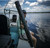 Toadfish Elite Stowaway Travel Rod, man holding compact fishing rod on a boat by the water under a cloudy sky.