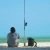 TOADFISH Toadfish Surf Combo, a person sitting on the beach with fishing gear and a fishing rod pointed toward the ocean. 