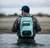 Toadfish Sealed Waterproof Backpack & Sling, a person wearing a blue cap and camouflage jacket standing in water with a Toadfish waterproof backpack showing on their back.