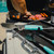 Toadfish Ultimate Grill Tongs, stainless steel grilling tongs with turquoise silicone handles resting on a grill table near a barbecue with cooking meat and sausages, outdoor setting.