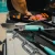 Toadfish Ultimate Grill Tongs, stainless steel grill tongs with teal silicone grips resting on a barbecue grill with food cooking in the background.