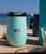 TOADFISH Toadfish Smartgrip Cups, mint green insulated travel cup with a black lid displayed on a wooden beach table by the ocean