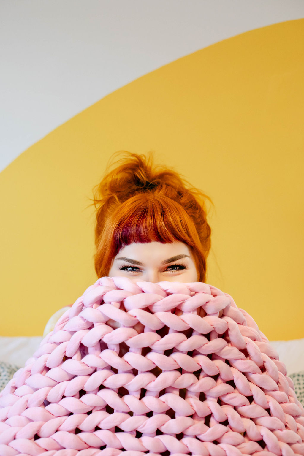 A blonde woman with a pink t-shirt, sitting on a white chair with a pink weighted blanket draped over her.