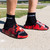 A close-up lifestyle shot of a model's feet wearing black socks and red and black camouflage CRNR foam slide sandals, standing outdoors on a concrete surface.