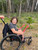 A smiling child with glasses sits in their GRIT Freedom Chair on a trail through a wooded area.