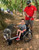 A young boy smiles as he pets a pot-bellied pig while sitting in his GRIT Junior all-terrain wheelchair. An adult man in a red t-shirt stands behind him, holding the wheelchair's push handles. They are outdoors in a wooded area with dirt ground, likely at a farm or petting zoo.