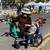 Smokey Bear poses with two young boys at an outdoor event. The boy on the right is seated in a GRIT Junior wheelchair, while the boy on the left stands wearing a green outfit. All three are pointing excitedly toward the left side of the image. White tents and other event attendees are visible in the background.