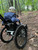 A young child wearing a blue patterned hoodie and a baseball cap is seen from behind, navigating a wooded trail in a GRIT Junior all-terrain wheelchair. The trail is covered in leaves, twigs, and roots, with green trees lining the path ahead