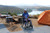 A young woman in a wheelchair smiles while sitting on Access Trax Mats at a scenic campsite overlooking a lake. The mats provide a stable and accessible surface on the uneven terrain.
