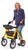 A smiling woman standing and holding the handles of a yellow Comodita Uno All-Terrain Rollator. The image demonstrates the rollator's sturdy frame, large pneumatic tires for rough terrain, and supportive backrest.