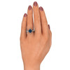 A hand with a light skin tone and manicured nails, painted a mauve color, displays a ring on the ring finger. The ring features a round alexandrite gemstone, displaying a mix of teal, blue, and purple hues. The gemstone is set in a 14k white gold band, flanked by triangular diamonds on either side, forming a snowflake design. The band is adorned with a row of smaller, round-cut diamonds, adding to the ring's sparkle. The background is white, allowing the ring to stand out.