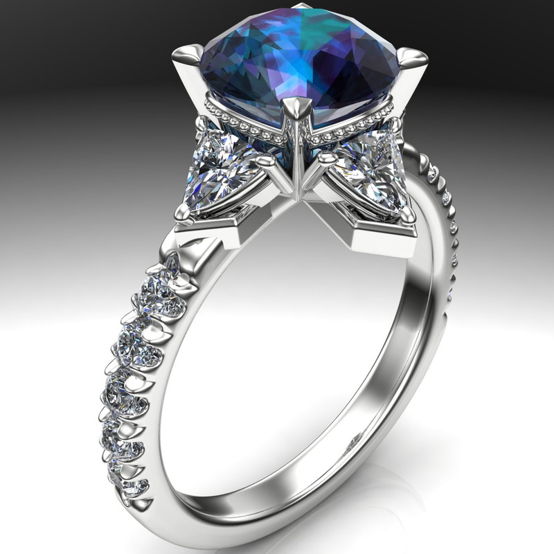 A close-up shot showcases a ring featuring a 2 carat round alexandrite gemstone, displaying a mix of teal, blue, and purple hues. The gemstone is set in a 14k white gold band, flanked by triangular diamonds on either side, forming a snowflake design. The band is adorned with a row of smaller, round-cut diamonds, adding to the ring's sparkle. The background is a gradient of gray, transitioning from dark to light, with the ring's reflection visible on the lighter surface.
