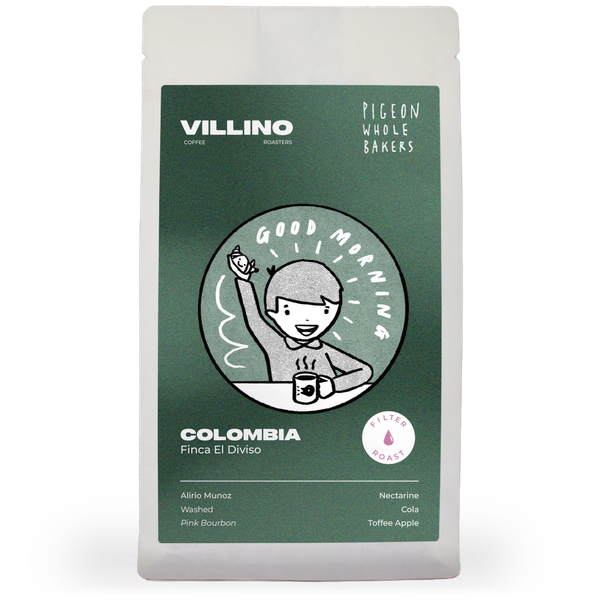 A bag of Single Origin Coffee on a white background. The dark green label features a light sage circle in the centre, with a cheery cartoon person with a coffee waving. In white text, it reads 'Pigeon Whole Bakers x Villino' and 'Colombia Finca El Diviso'.