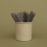 A pack of 16 bulk 10-inch UK-made straight dinner candles in charcoal grey, arranged in a neutral cream stoneware pot against a soft olive green background.