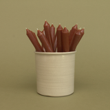 A pack of 16 bulk 10-inch UK-made straight dinner candles in dark brown, arranged in a neutral cream stoneware pot against a soft olive green background.