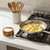 The RSVP Olive Wood Pinch Bowl, filled with salt, sits on a kitchen counter next to a stovetop, beside a stainless steel frypan filled with scrambled eggs.
