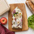 The Bees Wrap Plant-Based Sandwich Wrap in Meadow Magic print wraps a sandwich on a kitchen counter with an apple, lettuce, and bread.