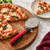 A Prep Solutions stainless steel pizza wheel sits next to a freshly cut pizza, with a slice on a nearby plate.