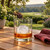 A golf ball-shaped ice cube made with the Tovolo Golf Ball Ice Molds in a rocks glass with bourbon on a teak patio table with a golf course in the background.