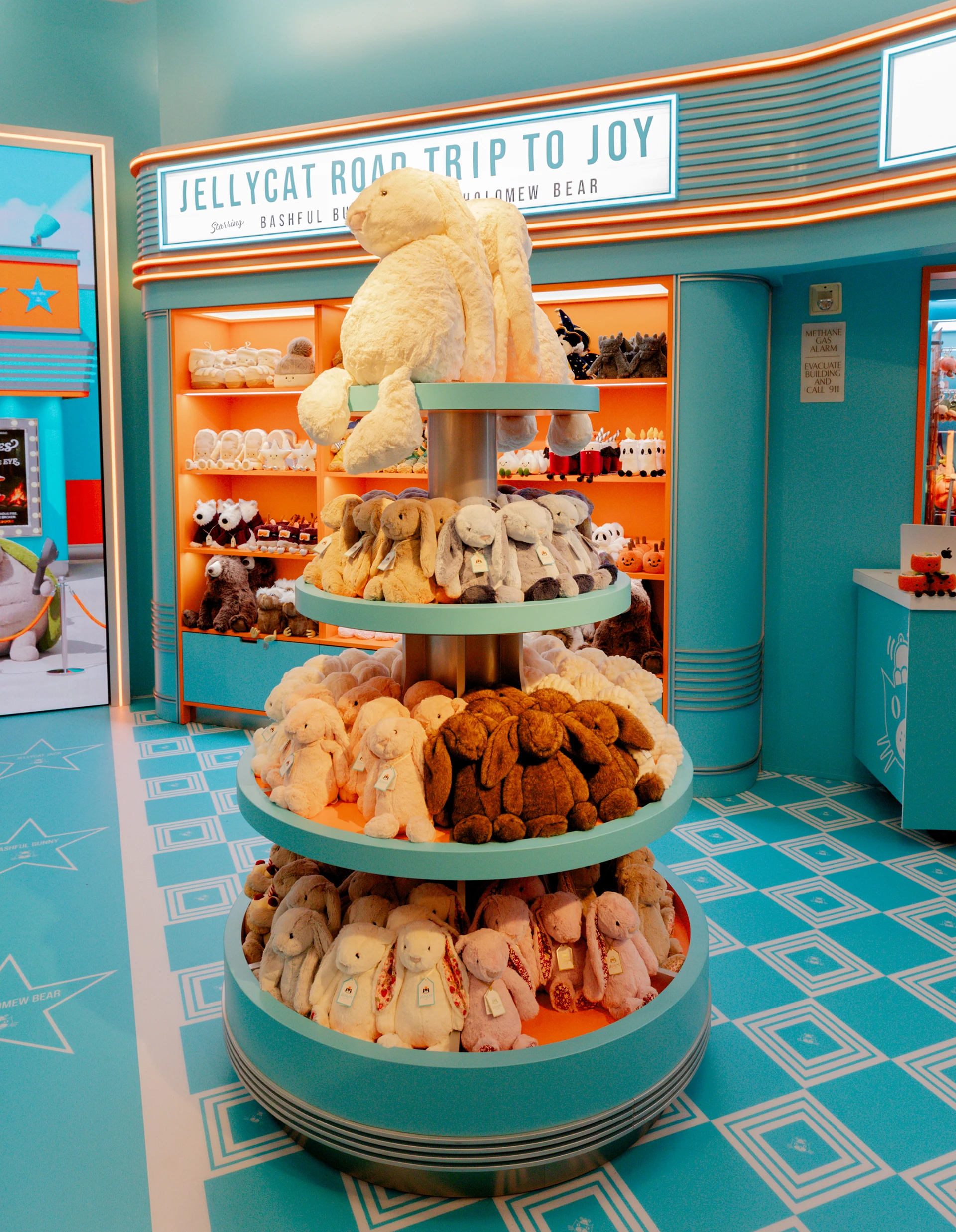 A large Jellycat Jack stands behind a retro cinema ticket counter surrounded by shelves of jellycats beneath movie marquee signs.