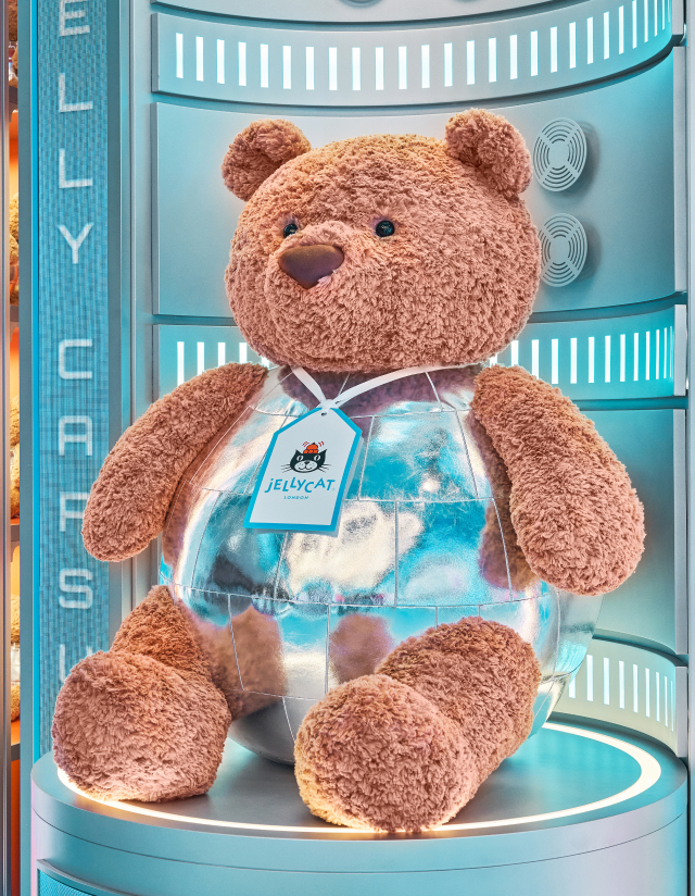 Bartholomew Bear sits in a capsule wearing a disco ball outfit beneath glowing Jellycat signage.