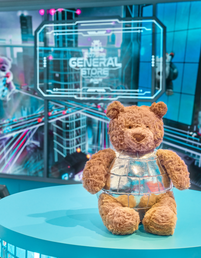 Bartholomew Bear wearing a disco ball outfit sits on a display table with a futuristic city screen behind.