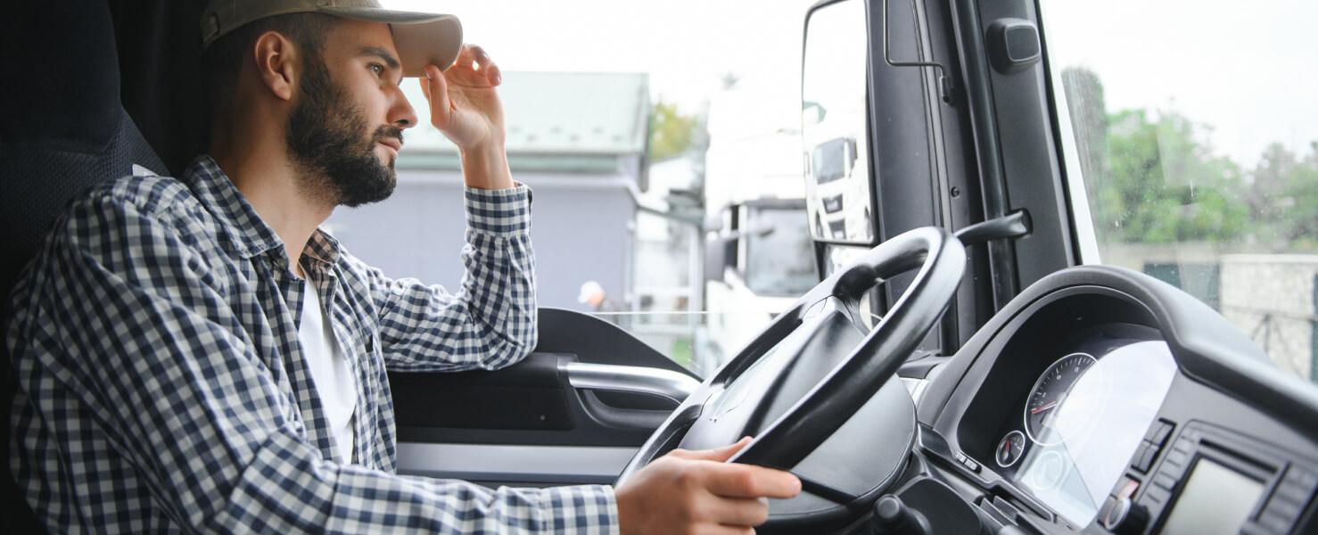 Truck driver wearing a cap and plaid shirt sitting behind the wheel of a commercial vehicle.