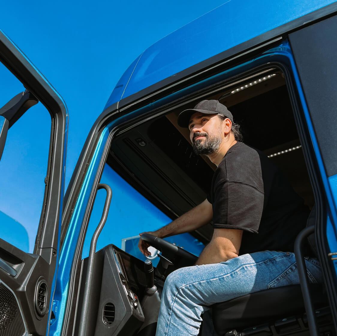 Truck driver in a black cap sitting in the cab of a blue semi-truck with the door open.