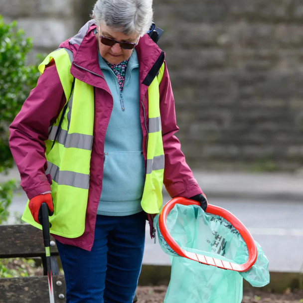 A lady wearing a hi-vis vest holds the handi-hoop fitted with green liner in one hand and a litter picker in the other