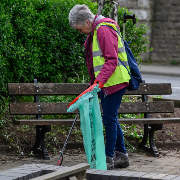 A lady wearing a hi-vis vest holds the handi-hoop fitted with green liner in one hand and a litter picker in the other