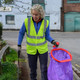 A lady wearing a hi-vis vest faces the camera while holding the handi-hoop fitted with a purple liner in one hand and a litter picker in the other