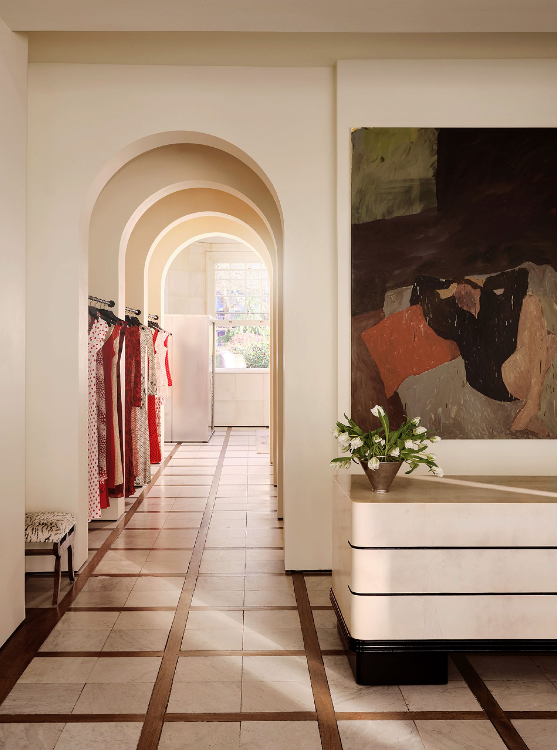 Enfilade of arched doorways past rails of dresses, with a painting above a stone chest of drawers