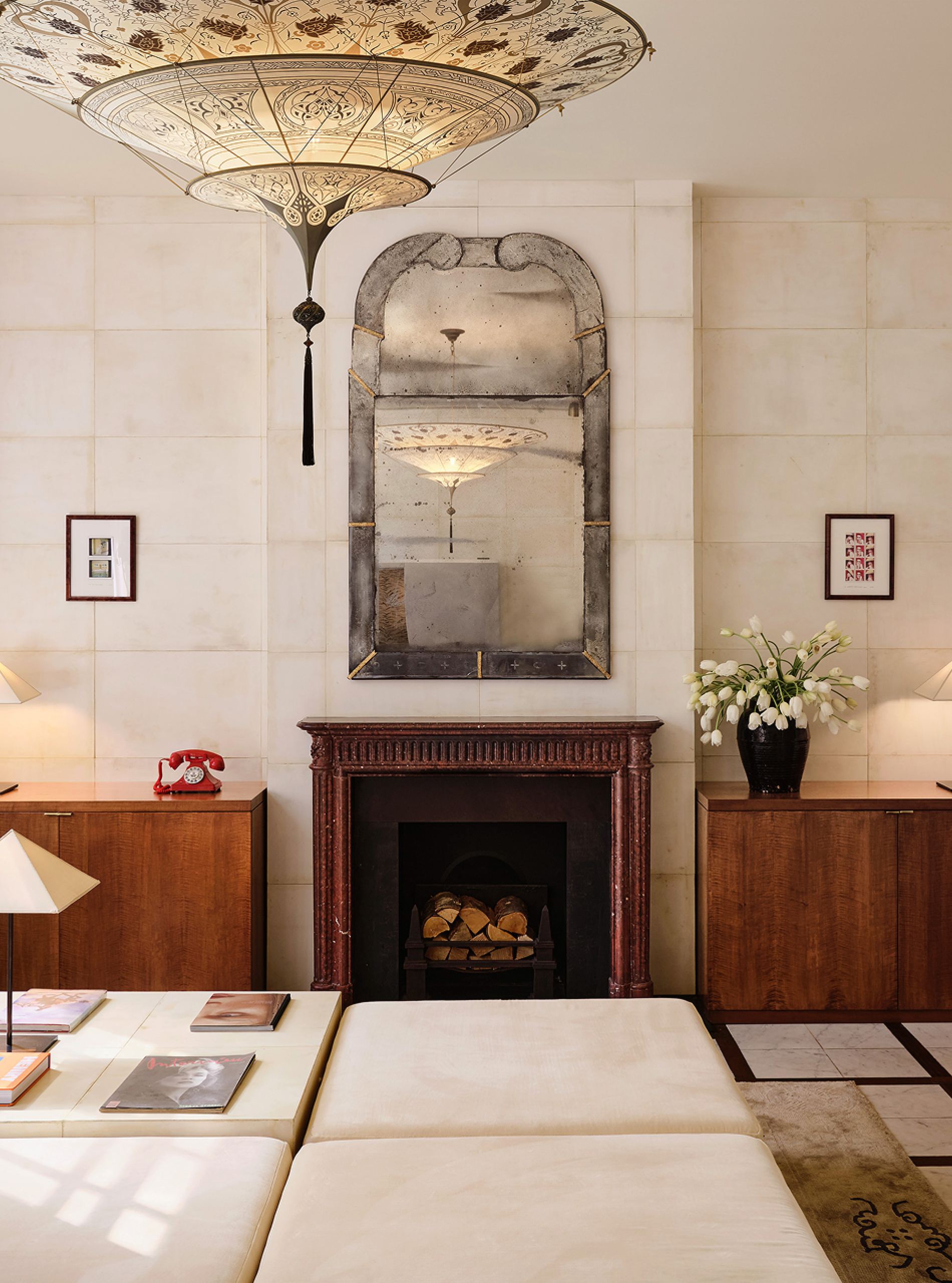 A sitting room with a dark wood fireplace, an arched mirror, and a painted paper lantern
