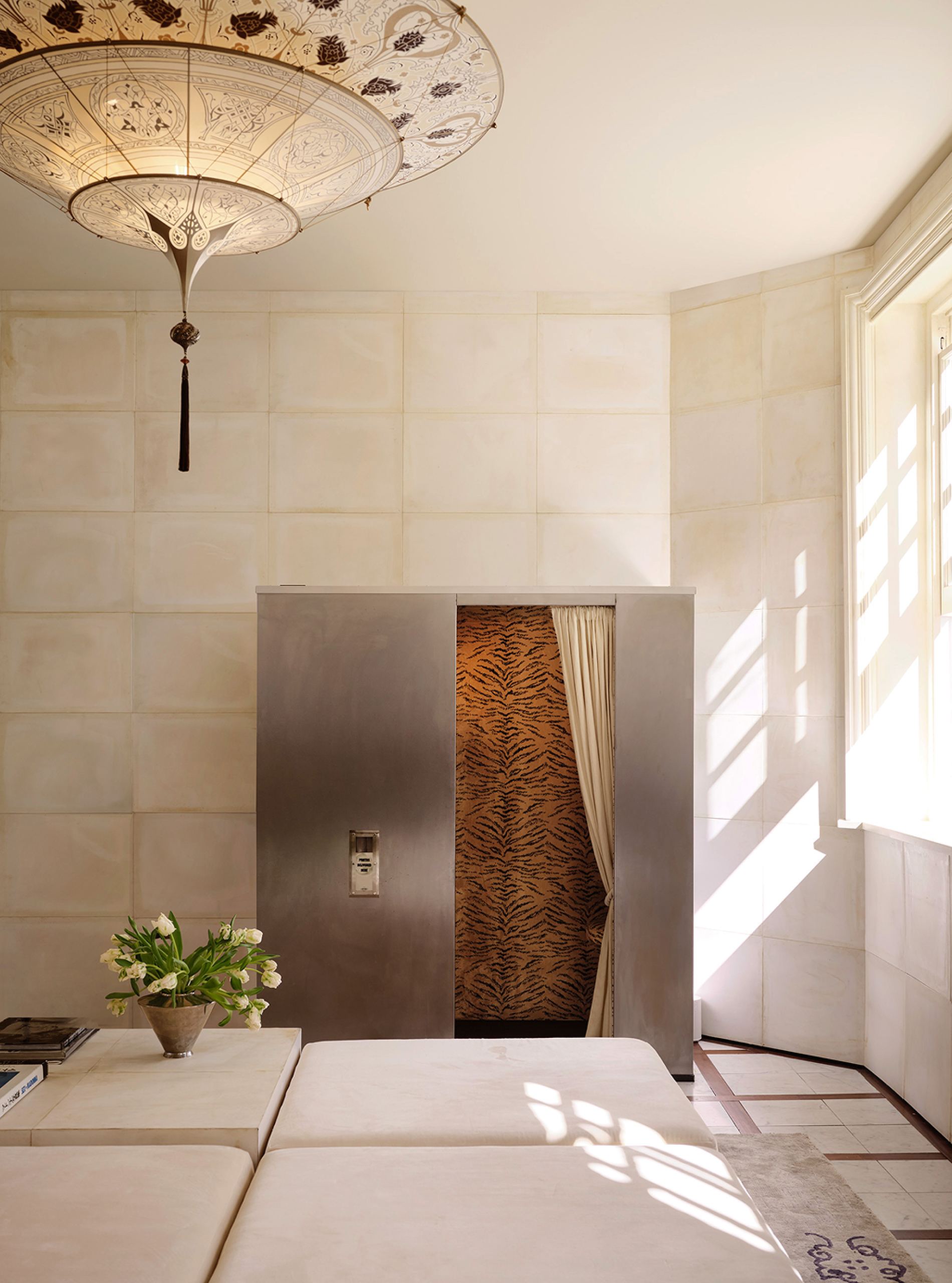 A metal photobooth with its curtain open, set into a parchment-tiled wall beside a sunlit window
