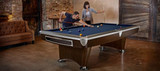Brunswick Billiards Brunswick Gold Crown VI Slate Pool Table | Skyline Walnut Finish, showcasing a dark wooden finish, with two people playing pool in a cozy, rustic indoor setting.