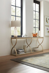 Hooker Furniture Elixir Console Table, elegant cream-colored wooden console table with curved legs, placed against a white wall with large window panels in the background, decorated with a lamp, glassware, a bowl of fruit, and decorative items in a modern interior setting.