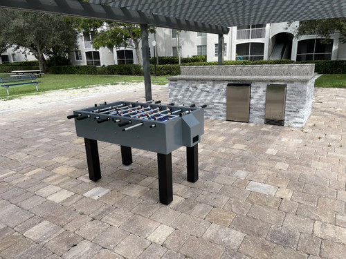 Sawyer Twain Outdoor Foosball, a modern outdoor foosball table with gray and black design placed on a brick patio in a park setting.