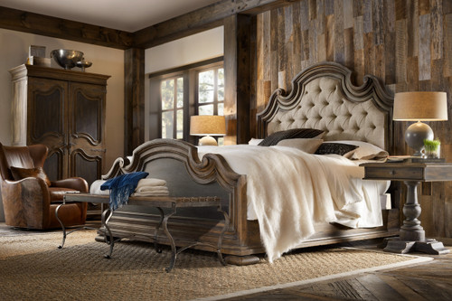 Hooker Furniture Fair Oaks King Upholstered Bed, rustic bedroom with tufted beige headboard, wood bed frame, matching bedside tables, lamps, leather armchair, wooden wardrobe, and natural lighting.