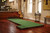 Brunswick Billiards Brunswick MacDonald 12' Indoor Putting Green - Espresso Finish, in a cozy living room setting with wooden flooring, guitars, and decorative wall art.
