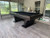 Plank & Hide Co Paxton Pool Table by Plank & Hide - Tobacco, a sleek black pool table with a modern design in a well-lit game room with large windows and wooden flooring.