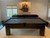 Plank & Hide Co Elias Slate Pool Table with Storage Drawer, a dark wooden pool table with black playing surface and a storage drawer, situated in a cozy room with white walls and windows.