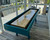 Venture Shuffleboard Mason Sport Shuffleboard Table by Venture, modern indoor shuffleboard table with a wooden playing surface and teal frame, located in a bright room with large windows and wooden flooring.