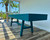 Venture Shuffleboard Mason Sport Shuffleboard Table by Venture, modern blue shuffleboard table in a sunlit room with large windows and palm trees outside.
