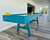 Venture Shuffleboard Mason Sport Shuffleboard Table by Venture, modern blue shuffleboard table in a bright room with wood flooring and large windows.