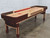 Venture Shuffleboard Grand Deluxe Bank Shot 9' Shuffleboard Table, made in USA, featuring a wooden playing surface with scoring zones and a red puck, set on a sturdy wooden frame with a corner bumper for gameplay.
