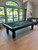 Doc & Holliday Zurich Pool Table by Doc & Holliday with Black and Custom Finishes, placed in a bright room with large windows overlooking a neighborhood.